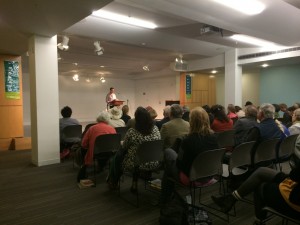 Annaliese Jakimides reads her poetry in front of a seated crowd. 