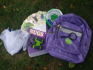 Purple backpack on the grass with the following contents surrounding it: butterfly net, binoculars, magnifying glass, two way bug viewer, insect habitat, clipboard case.