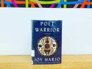 A hardcover copy of the book "Poet Warrior" stands on a wooden surface in front of a white wall. A mural with colorful curving lines is on the wall above the book. On the book cover below the title there is an image of beadwork art in a circle pattern with a beadwork spider in the middle.
