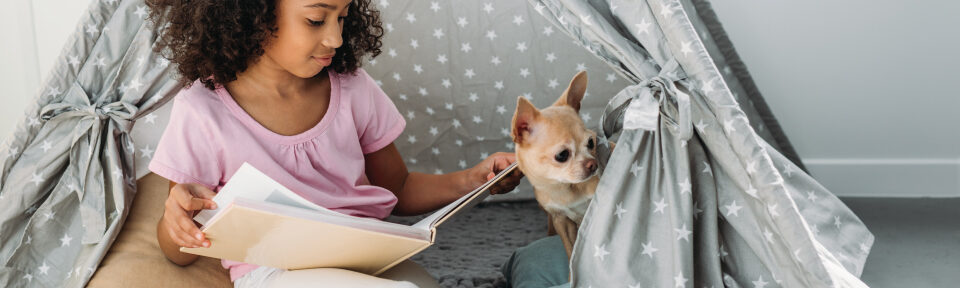 Girl with dark skin and curly hair reading a book to her pet dog