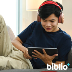 Boy watches a video on his tablet while lounging on a couch. biblio+ icon is in the bottom right corner.