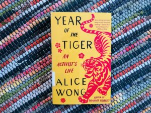 A yellow and red copy of Year of the Tiger: An Activist's Life by Alice Wong with a red tiger on the cover sits on a multicolor-striped background