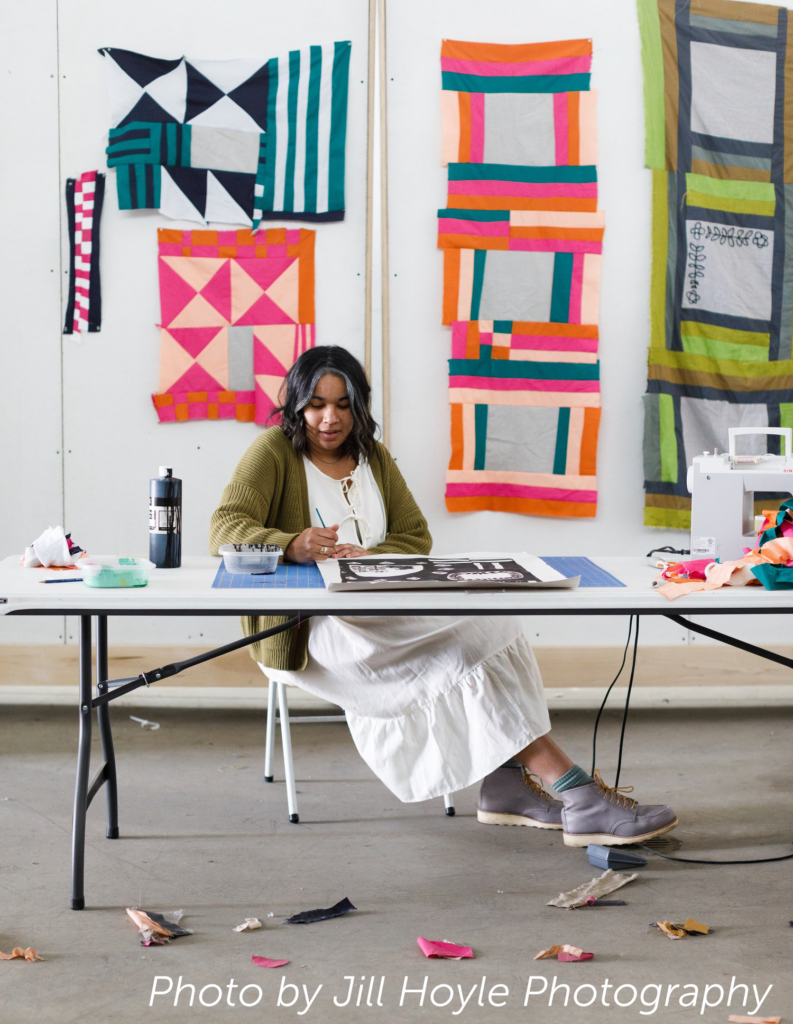 Rachel Adams sits at a table creating a painting in front of various textile artworks.
