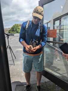 Researcher, Dr. Purlin, holds a herring gull chick on the roof of the Downtown Library. Dr. Purlin wears a backpack with tagging equipment, binoculars, and a scope can be seen on a tripod behind him.