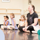 Four children sit with a ballet instructor at a pre-ballet class